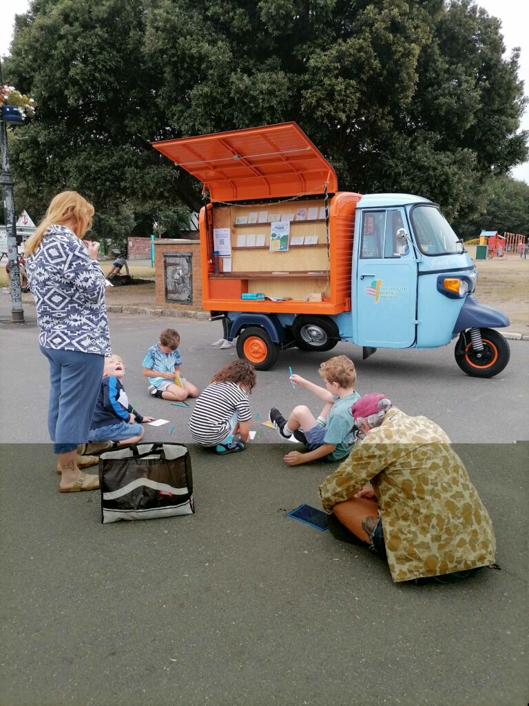 People drawing outside in the park with a tuktuk and trees in the background